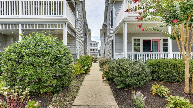 A narrow pathway flanked by neatly trimmed hedges and small plants, leading to a row of two-story buildings with white railings and a red door. - Ocean City Home 1 Block to Beach & Boardwalk! (Ocean City)