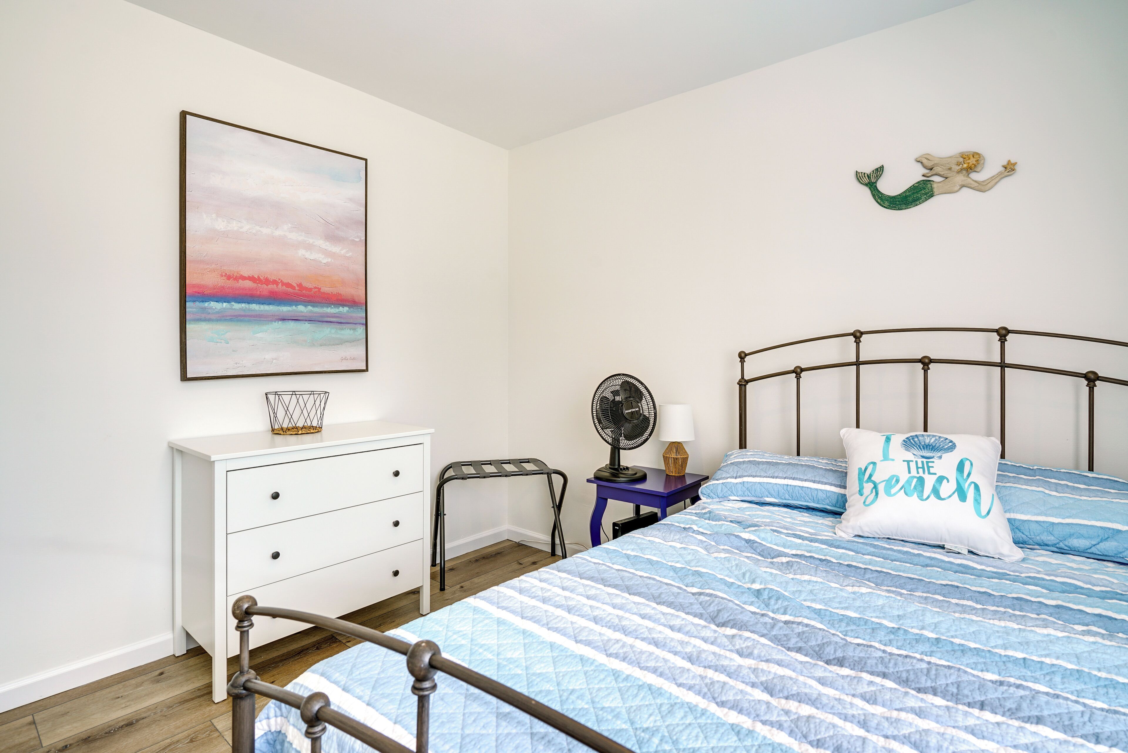 Une chambre à coucher comprenant un lit, une table de chevet, une commode, un ventilateur fixé au mur et un coussin à motifs de plage.