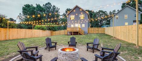 A backyard with a stone fire pit surrounded by Adirondack chairs, a wooden deck, and a house with string lights.