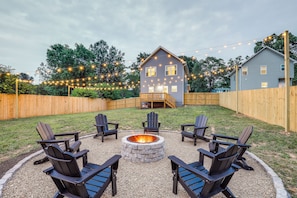 A backyard with a stone fire pit surrounded by Adirondack chairs, a wooden deck, and a house with string lights. - Getaway w/ Fire Pit: 3 Mi to River Arts District! (Asheville)