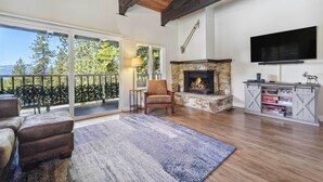 A spacious living room with a fireplace, a television mounted on the wall, and a balcony with a view of trees and mountains. - 10 Mi to Palisades Tahoe! Condo w/ Game Room (Tahoe City)