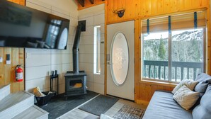 L’intérieur chaleureux d’un chalet avec une cheminée, un téléviseur fixé au mur et une grande fenêtre donnant sur la forêt.