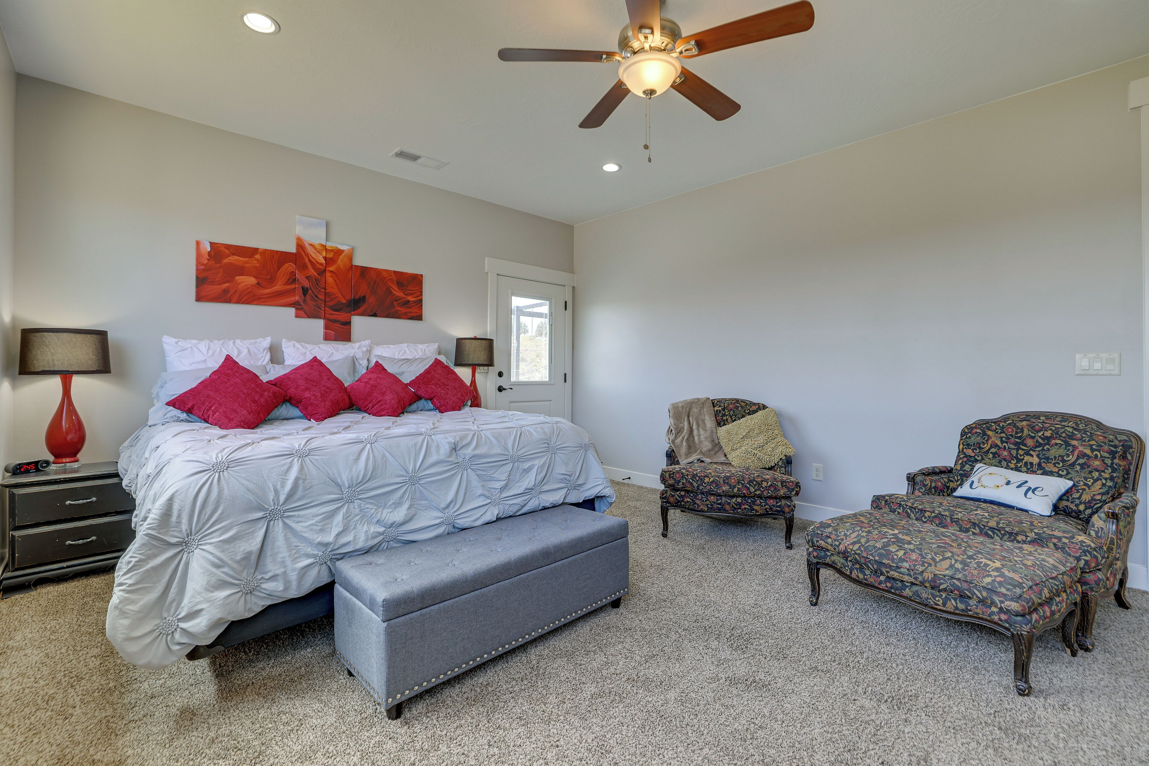 A bedroom with a bed, a bench, two chairs, a ceiling fan, and a door.