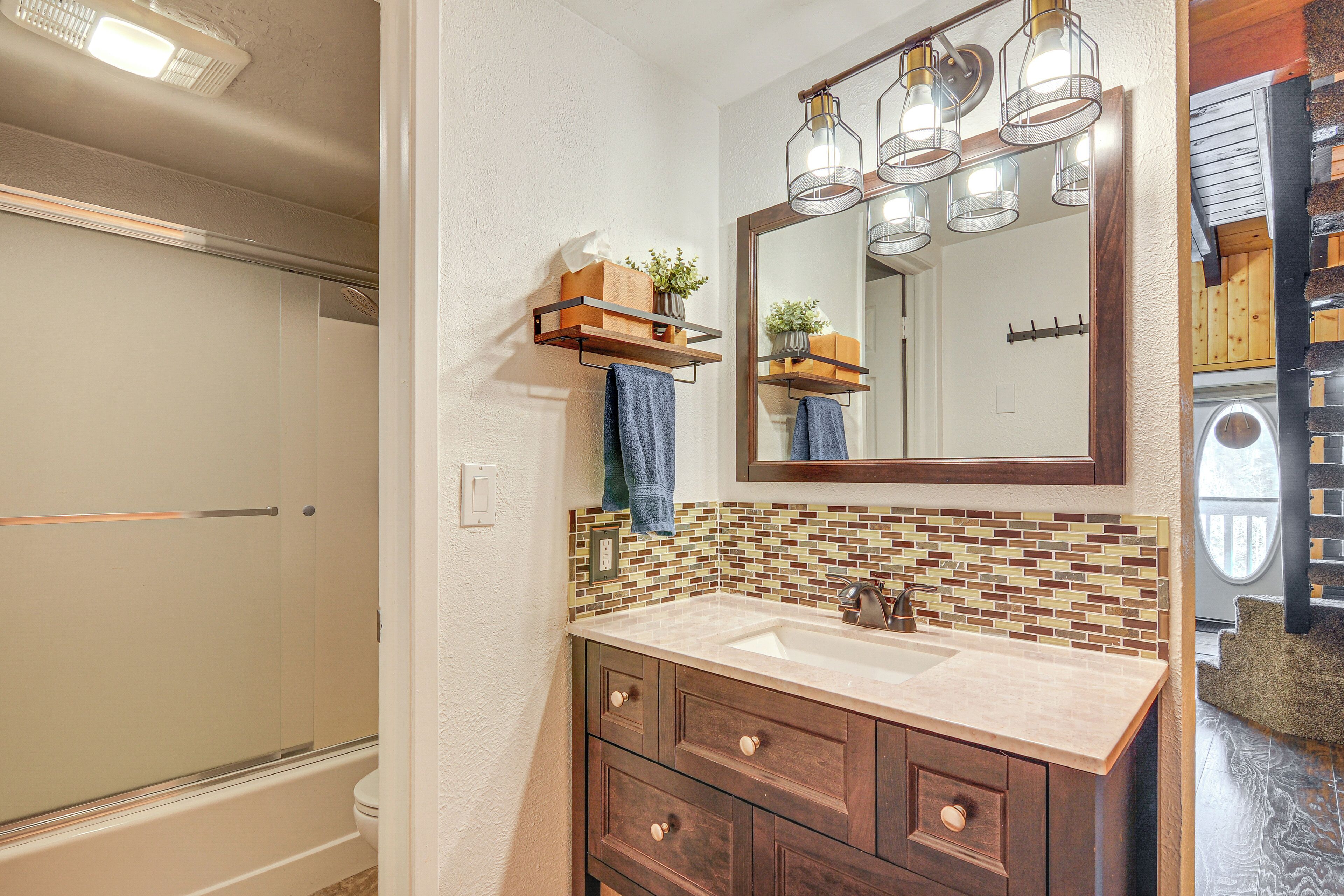 A bathroom with a glass shower enclosure, a beige sink with a wooden vanity, a mirror with pendant lights, and a tiled backsplash.