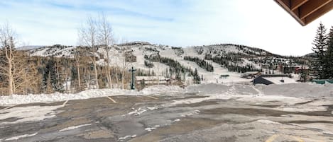 A parking lot with snow-covered ground and a view of a mountainous area with trees and buildings in the background.