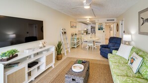 A living room with a flat-screen TV, a ceiling fan, a green patterned sofa, and a dining area in the background. - Ocean-view Key Colony Beach Condo w/ Pool Access! (Key Colony Beach)