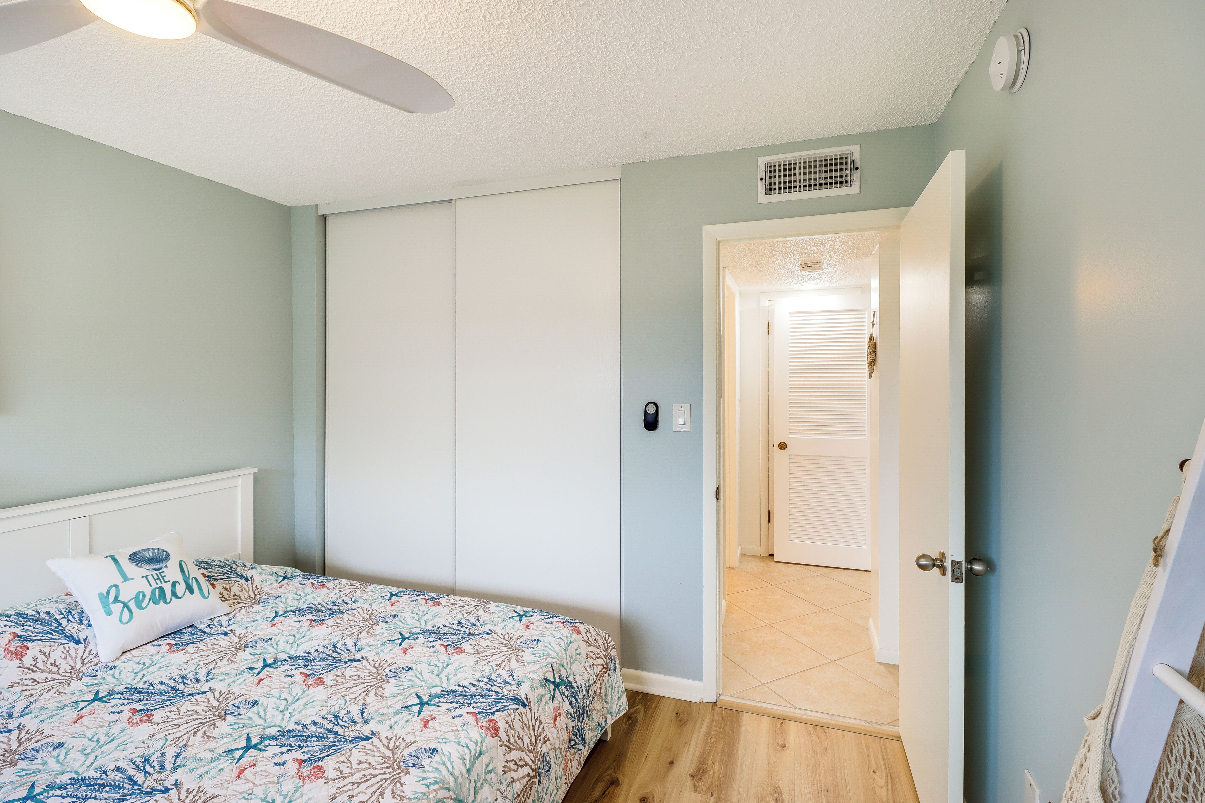 A bedroom with a bed, a ceiling fan, a door leading to another room, and a light switch on the wall.