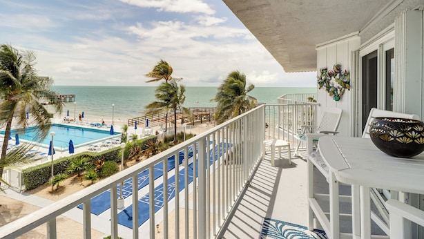 A balcony with a view of a pool and beach, white railings, and a decorative vase. - Ocean-view Key Colony Beach Condo w/ Pool Access! (Key Colony Beach)