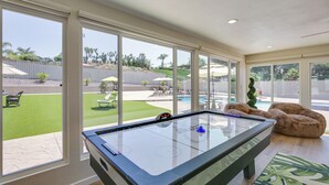 Ein Billardtisch in einem Raum mit großen Fenstern, die auf einen Garten mit Pool, Terrassenmöbeln und einem Zaun blicken.