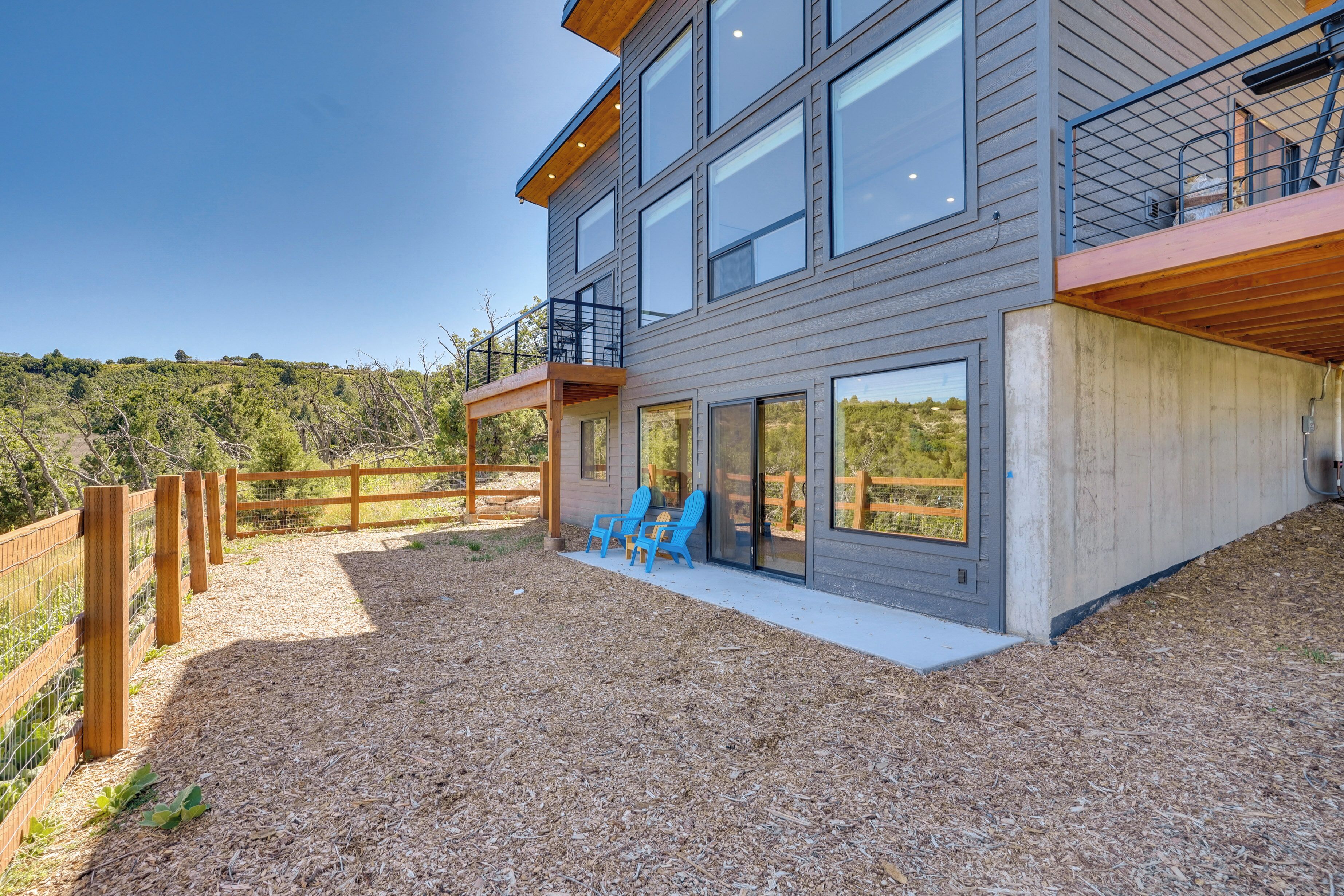 Une maison moderne à deux étages, dotée de grandes fenêtres, d’un balcon et d’un patio couvert avec des chaises bleues.