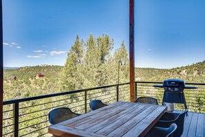 A wooden deck with a grill, chairs, and a table overlooking a hilly landscape with trees and buildings.