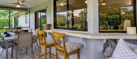 Ein überdachter Patio-Bereich mit Tisch und zwei Holzstühlen, einem Deckenventilator und großen Fenstern mit Blick auf Grünflächen.