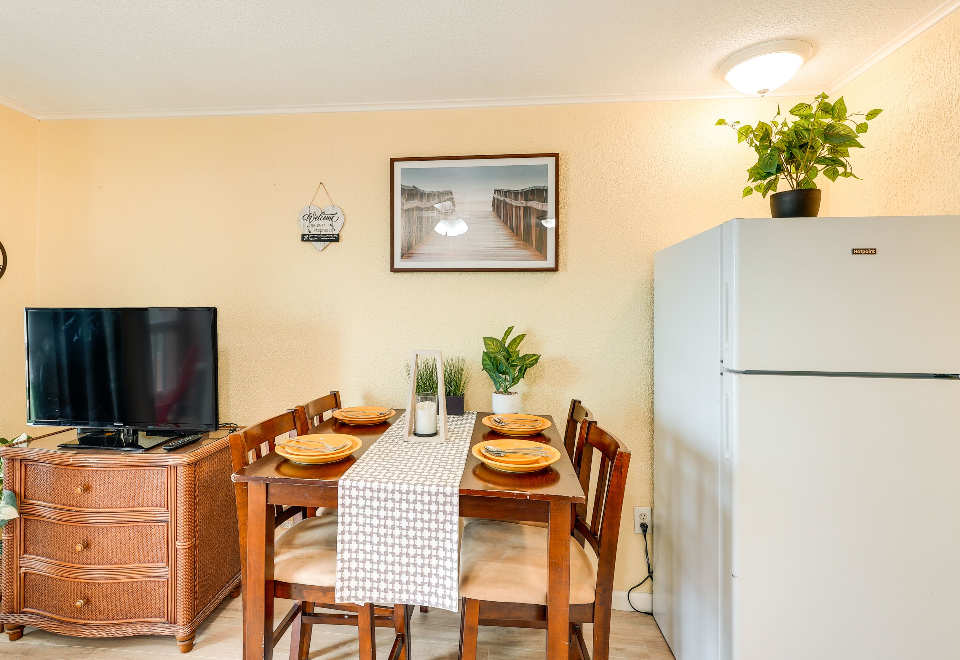 A dining area with a table set for four, a television on a cabinet, a refrigerator, and a plant on the counter.