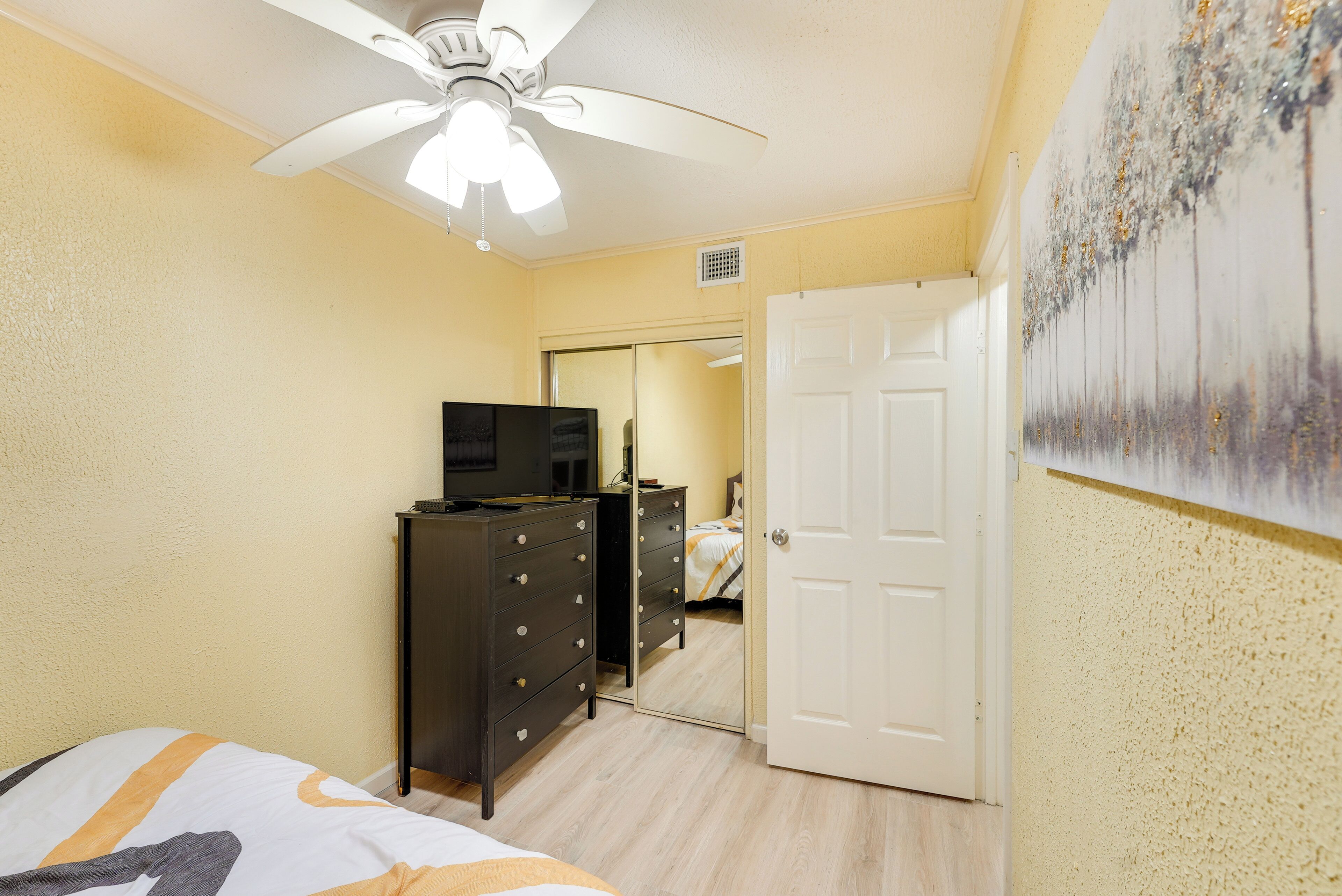 Un dormitorio con cama, cómoda, ventilador de techo, puerta y un cuadro en la pared.