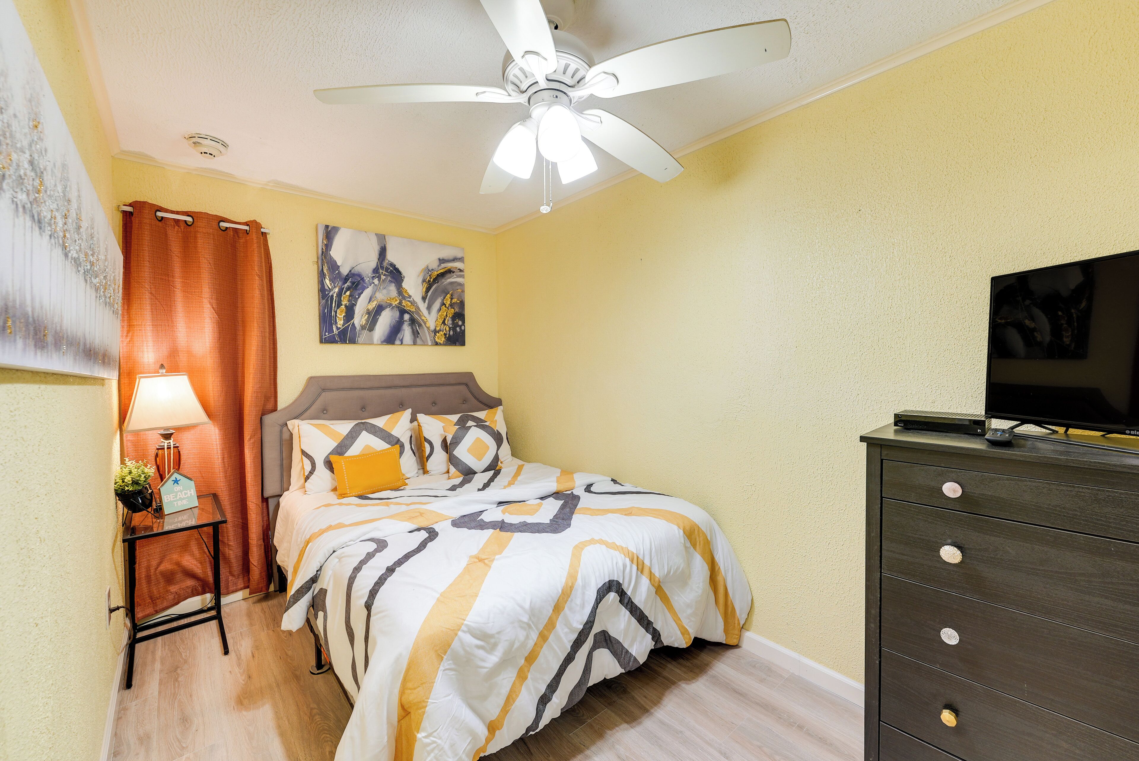 A bedroom with a bed, a nightstand, a dresser, a ceiling fan, and a painting on the wall.