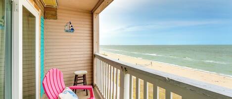 A balcony with a view of the beach, a wooden wall with a 'BEACH' sign, and two pink chairs.