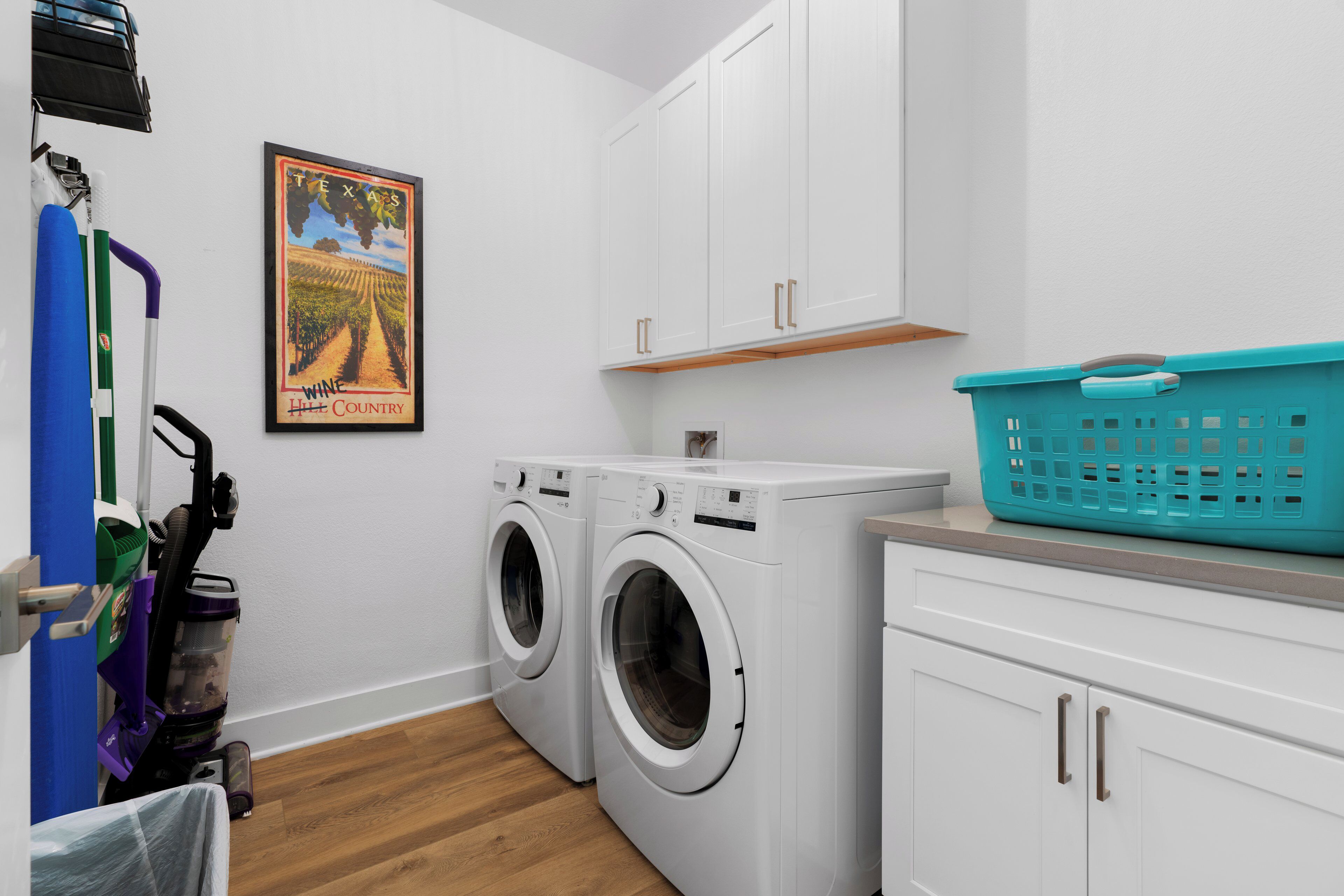 A laundry area with two front-loading washing machines, a blue laundry basket, and a poster of Texas wine country on the wall.