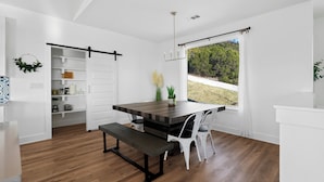 A modern kitchen with a wooden table, white chairs, and a view of greenery outside. - Lago Vista Home w/ Game Room & Outdoor Tub! (Leander)