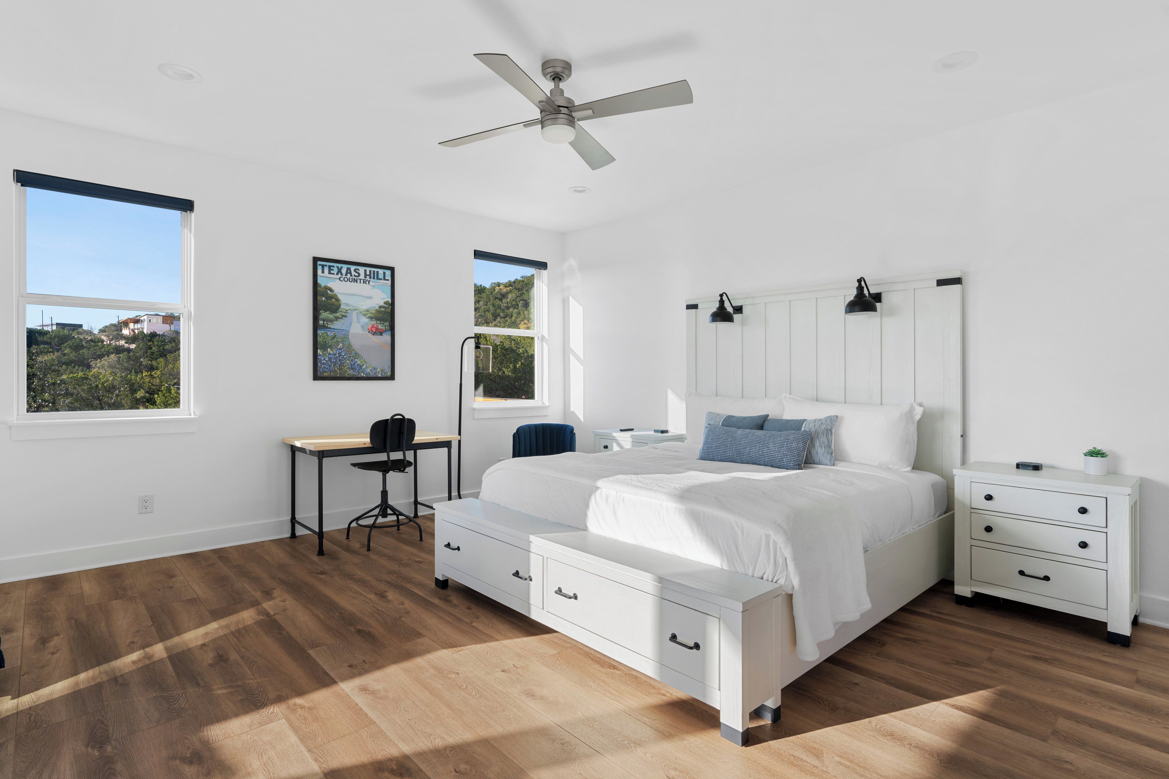 A bedroom with a large bed, a desk, and a chair, a ceiling fan, and a view of a hill.