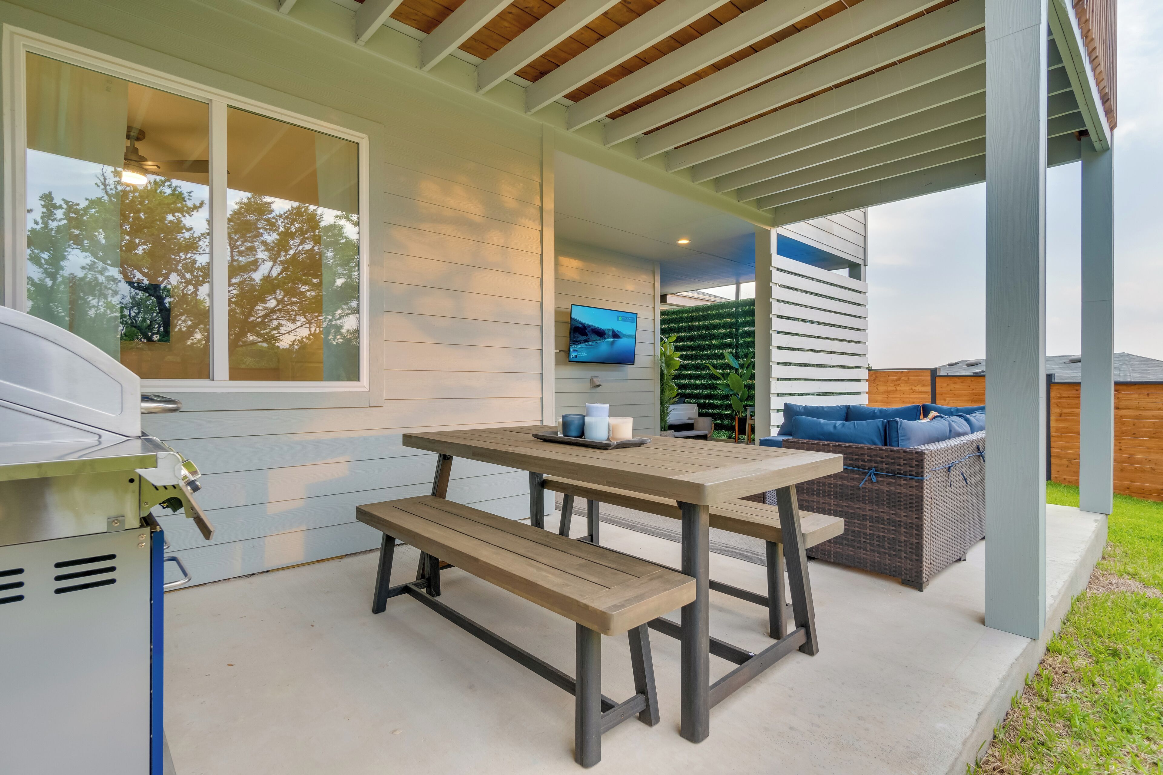 A patio with a wooden table and bench, a grill, and a TV mounted on the wall.