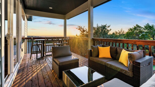 A covered outdoor deck with wicker furniture, a glass table, and a view of the sunset over a body of water. - Lago Vista Home w/ Game Room & Outdoor Tub! (Leander)