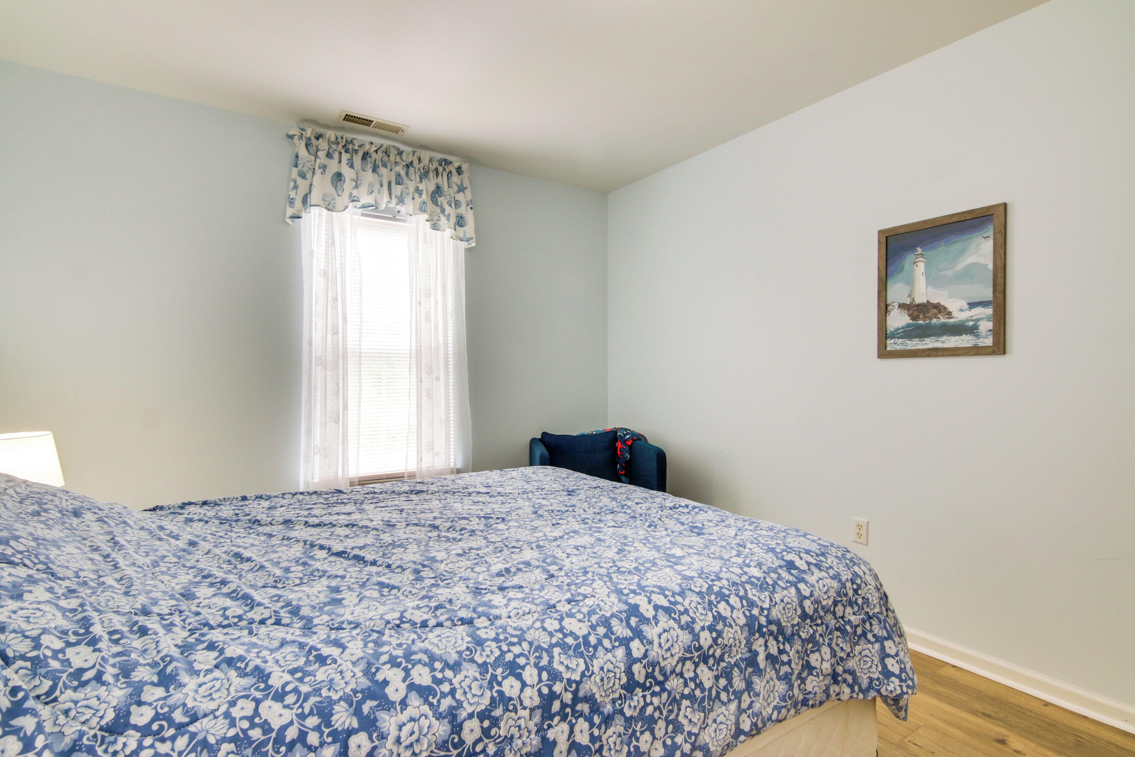 Un dormitorio con una cama grande, una ventana con cortinas translúcidas, un cuadro de un faro y una silla.