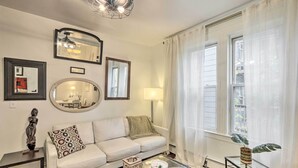 A living room with a white sofa, a glass coffee table, a lamp, and a large window with sheer curtains.