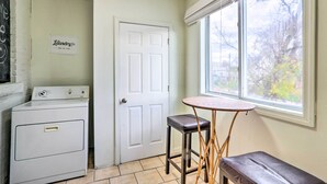 A laundry room with a washing machine, a dryer, a door with a sign, a small table with a stool, and a window with a view of trees.