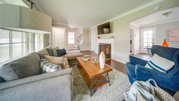 A spacious living room with a sofa, armchair, and a wooden coffee table. A fireplace is visible in the background, and there is a staircase leading to another level. - Vacation Rental Near Cleveland, Ohio: 6 Mi Away! (University Heights)