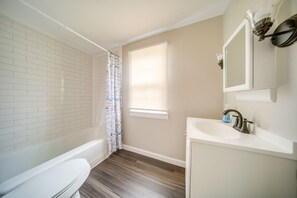 A bathroom with a white toilet, a bathtub, a sink with a mirror, and a window with blinds. - Vacation Rental Near Cleveland, Ohio: 6 Mi Away! (University Heights)