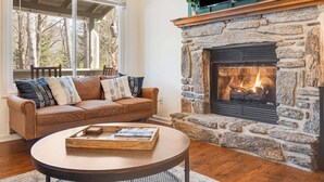 A cozy living room with a stone fireplace, a leather sofa with patterned cushions, and a round coffee table with magazines.