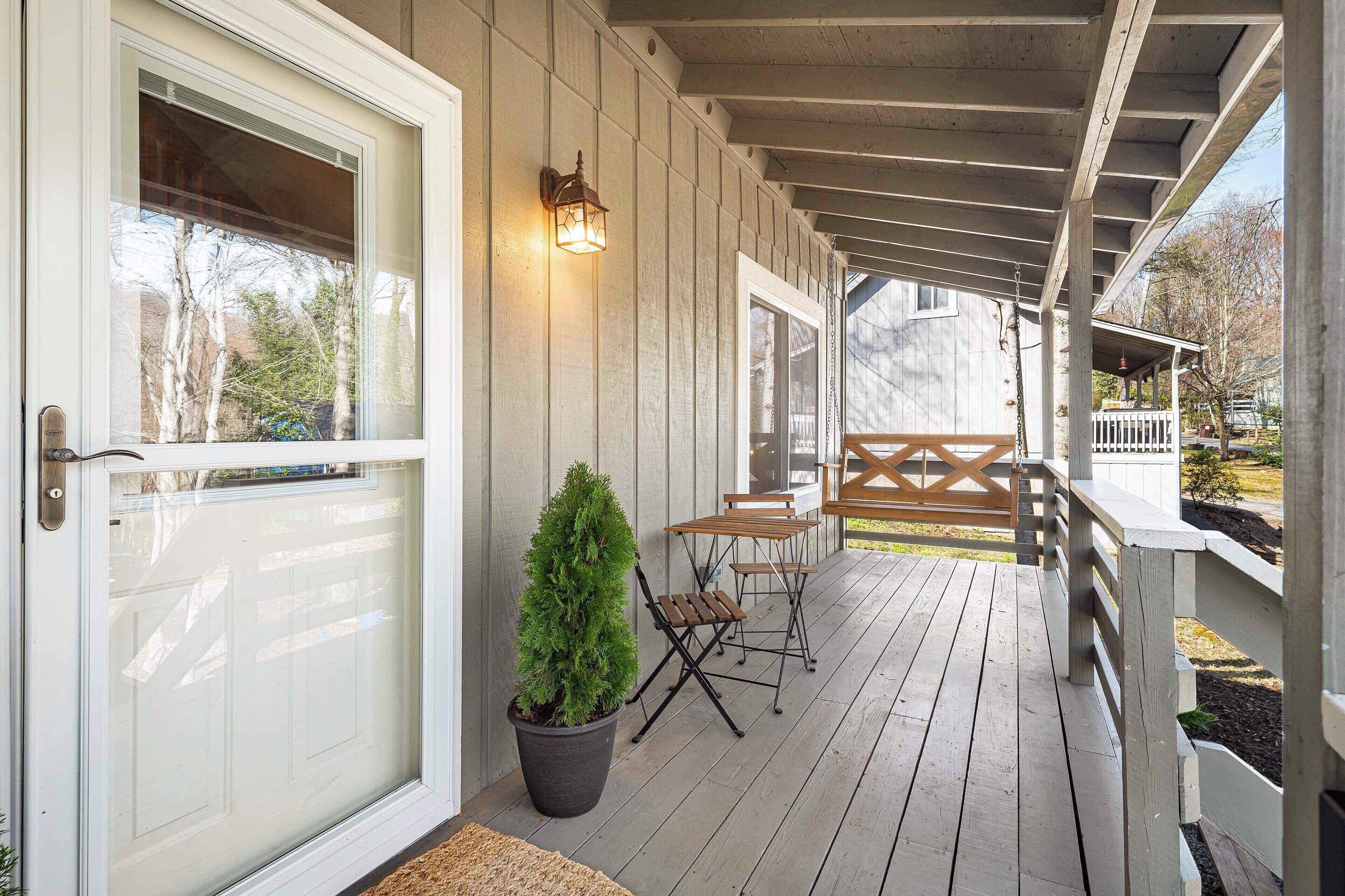 A covered porch with a wooden deck, a small table and chairs, a potted plant, and a wall-mounted light fixture.