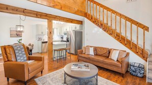 A spacious living area with a wooden staircase, leather sofas, and a kitchen in the background.