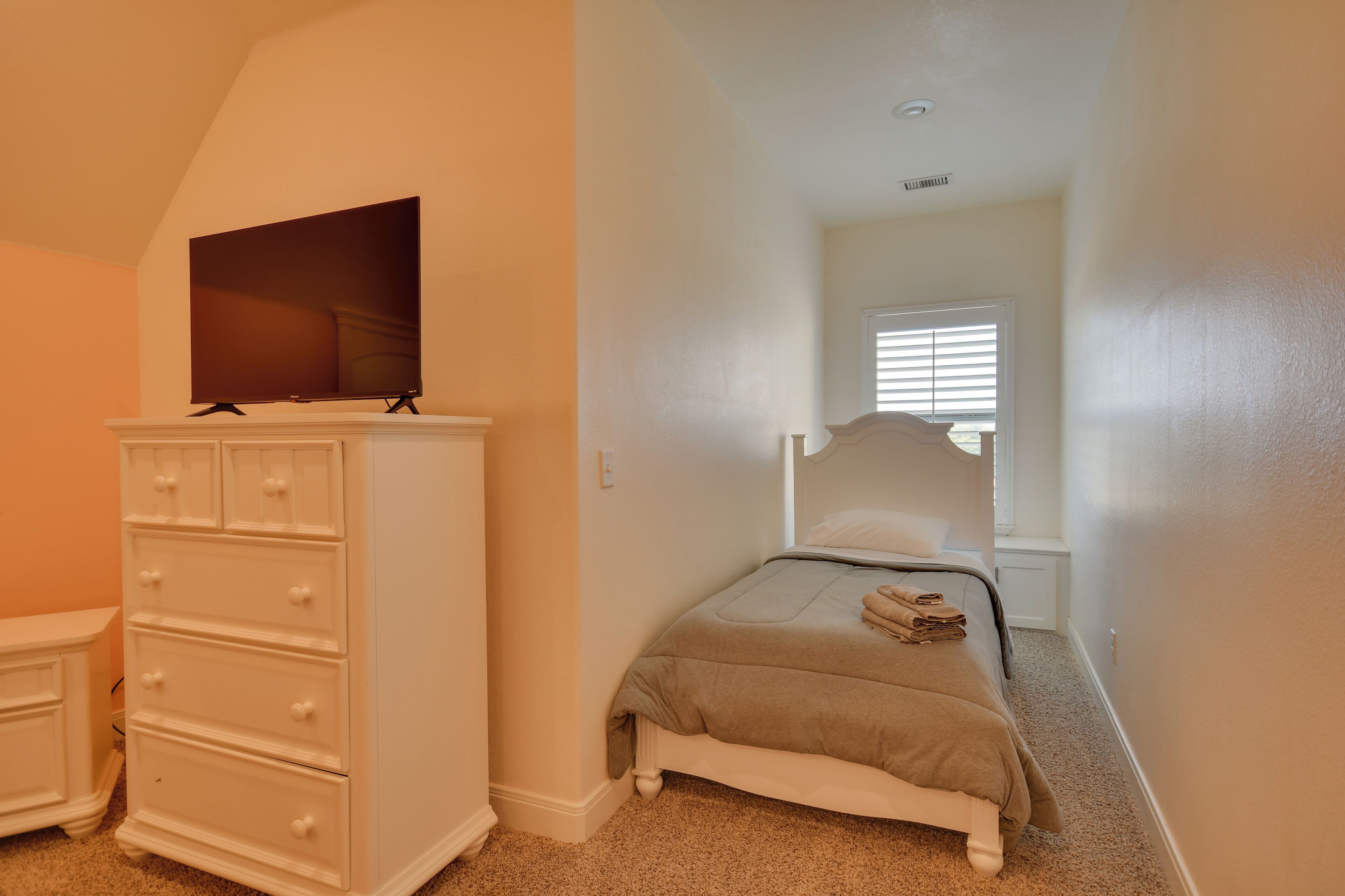 A bedroom with a bed, a dresser, a television on top of the dresser, and a window with blinds.