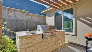 Eine Außenküche mit Gasgrill, eingebautem Spülbecken und Fenster mit Blick auf einen Garten.