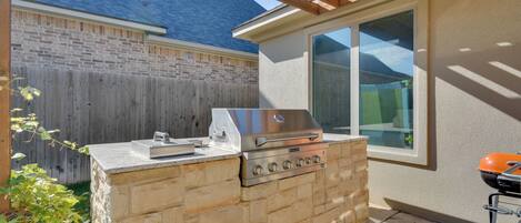 An outdoor kitchen with a gas grill, a built-in sink, and a window overlooking a backyard.