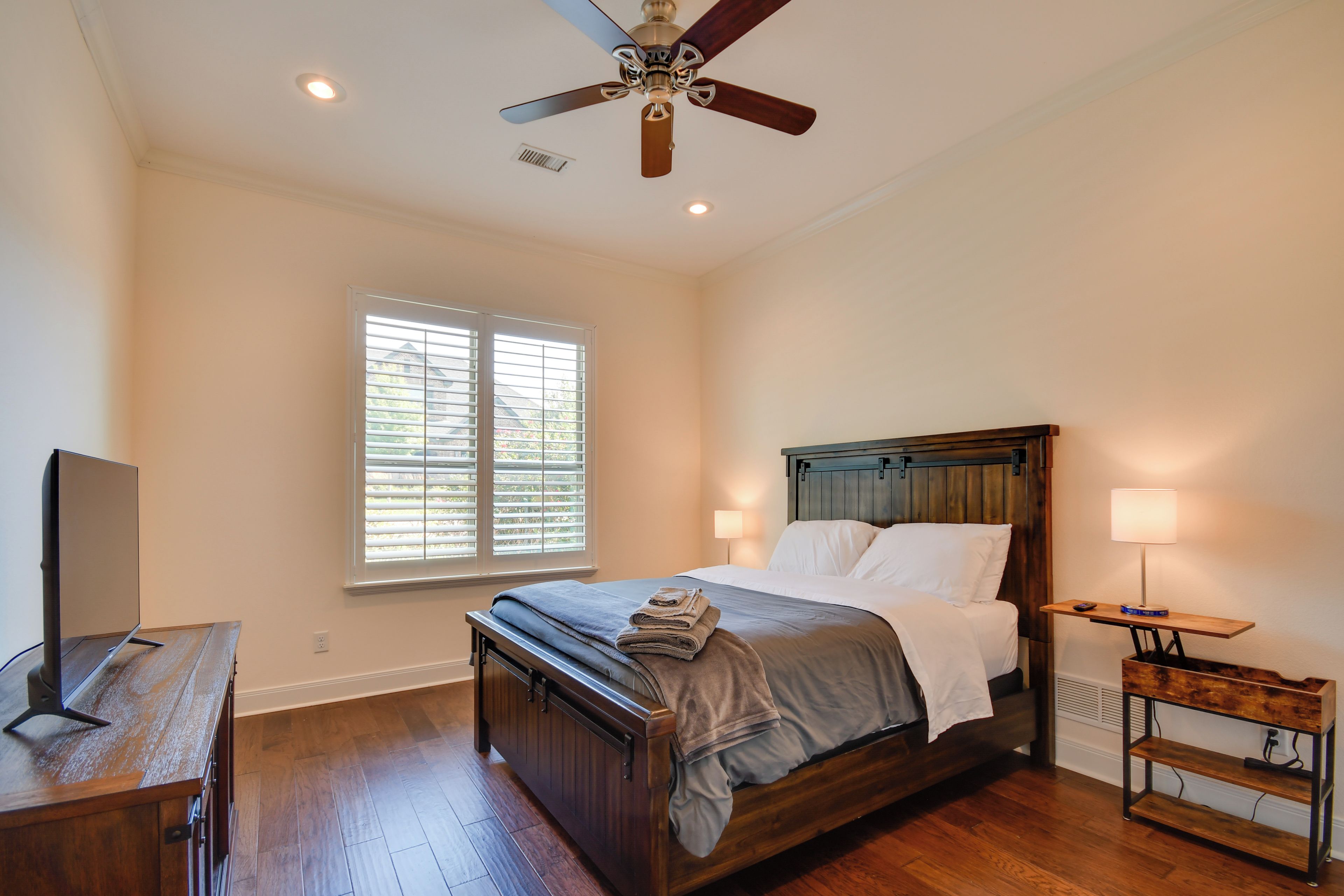 A bedroom with a wooden bed, a desk, a ceiling fan, and a window with blinds.