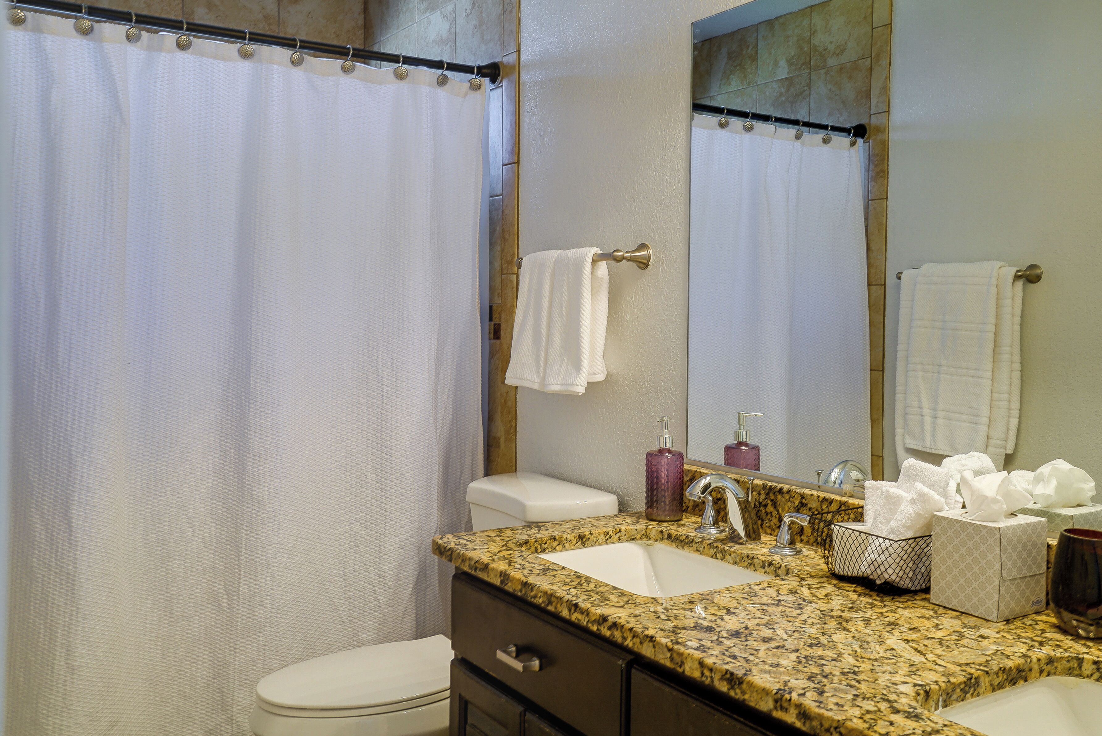 Une salle de bain équipée d’un rideau de douche, de toilettes, d’un lavabo avec un plan de travail en marbre, d’un miroir et d’un porte-serviettes.