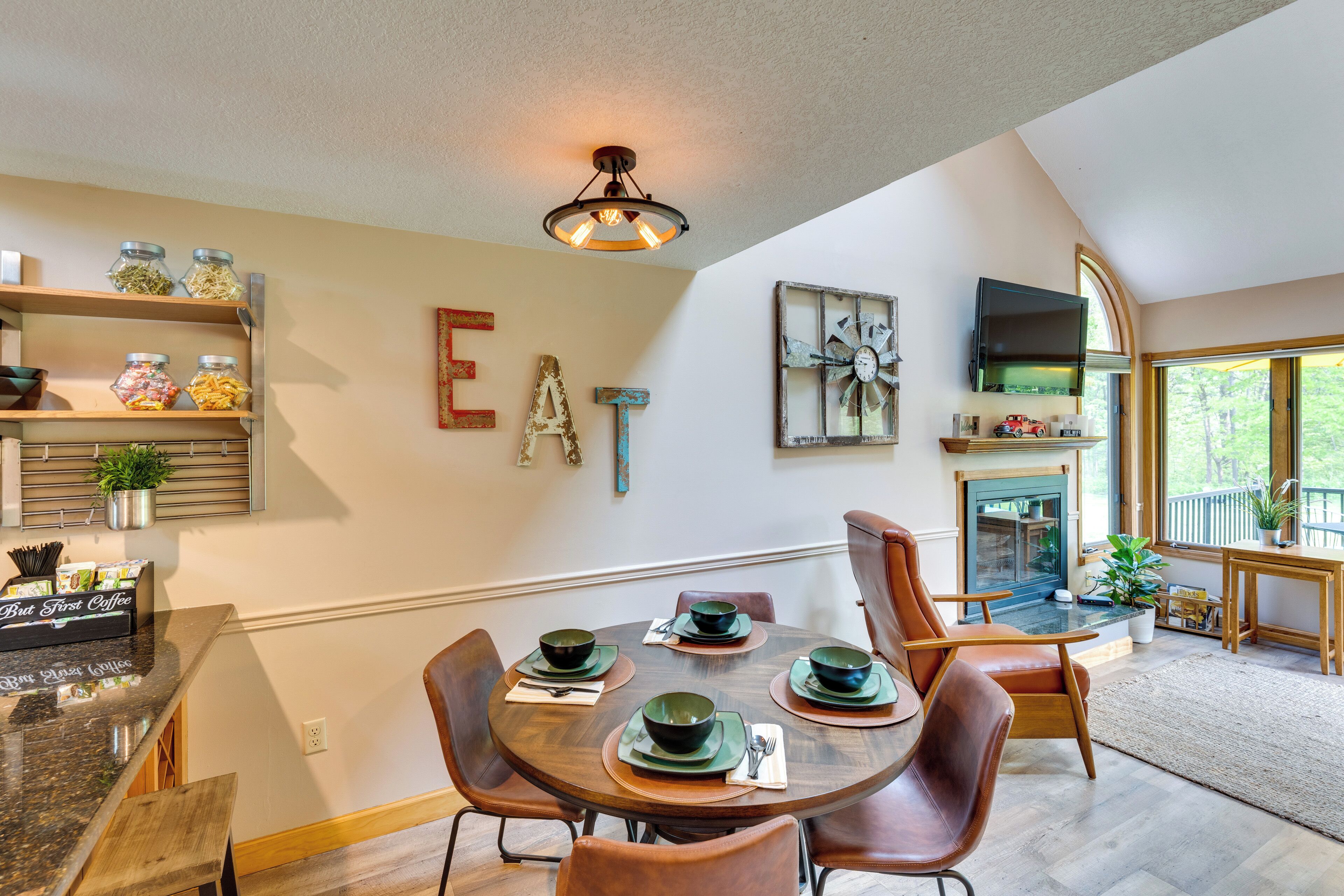 A dining area with a round table set for four, surrounded by leather chairs. There are shelves with jars and a But First Coffee sign. A fireplace with a TV above it is visible, and a large window offers a view of the outdoors.