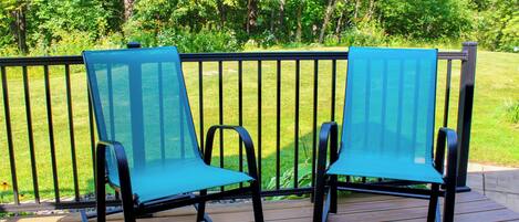 Two blue chairs on a wooden deck with a metal railing, surrounded by greenery.