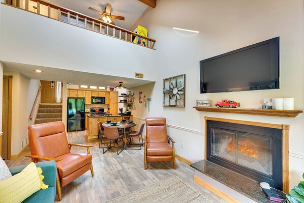 A spacious living room with a fireplace, a television mounted on the wall, and a kitchen area visible through an open doorway.