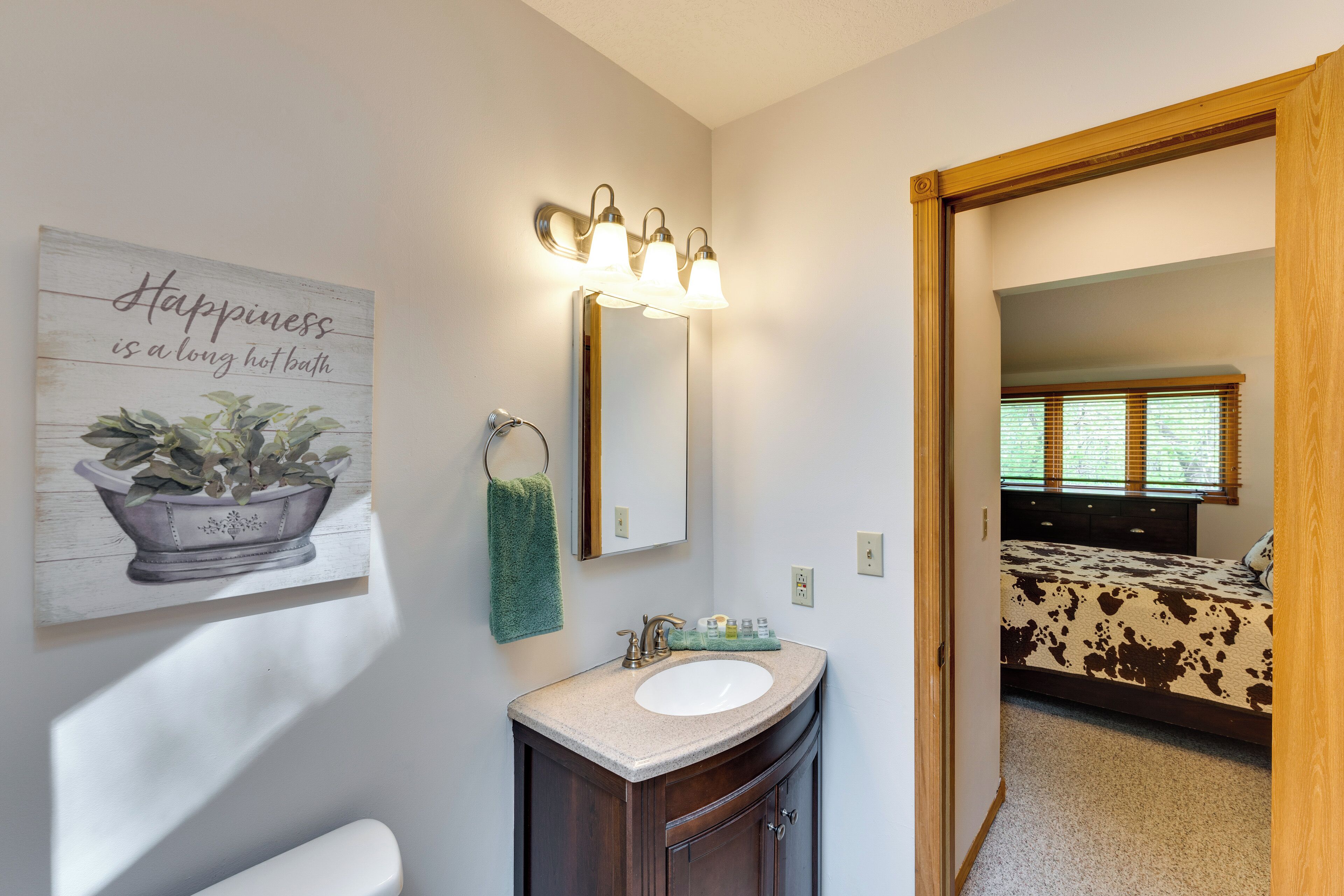 A bathroom with a sink, vanity, and a mirror. There is a wall decoration with a potted plant and a quote. A towel hangs on the wall.