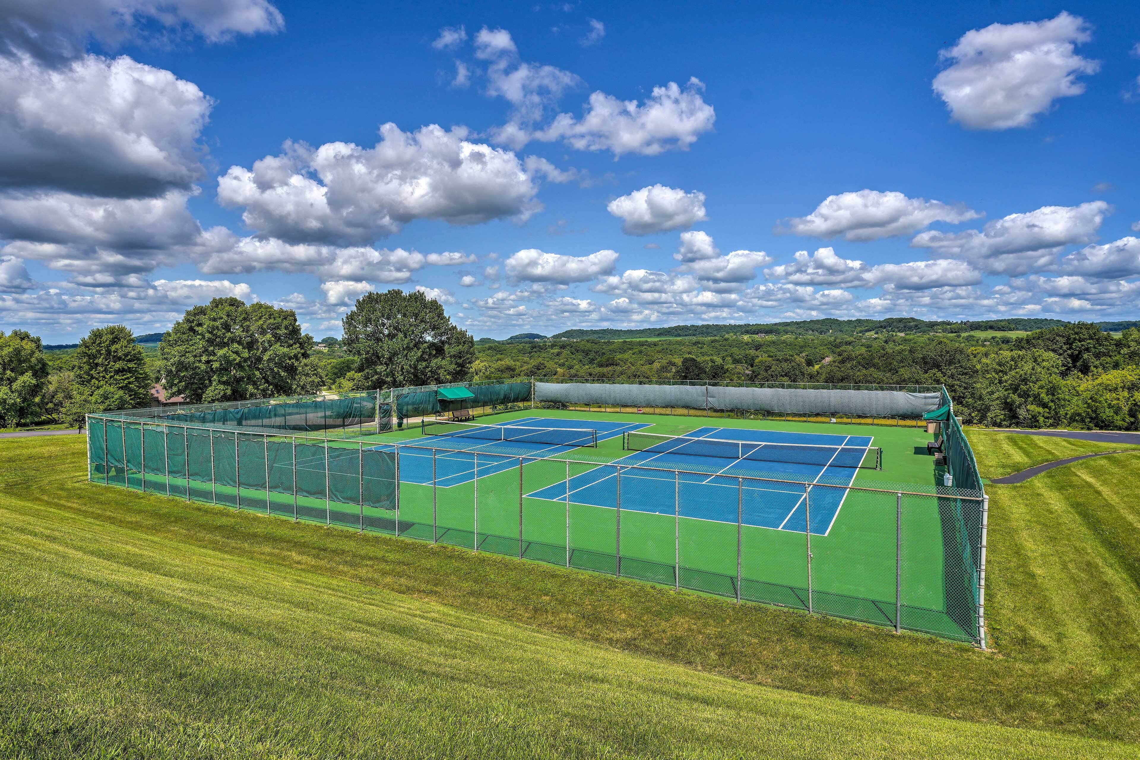 A tennis court with a green fence and net, surrounded by a grassy field and trees under a blue sky with clouds.