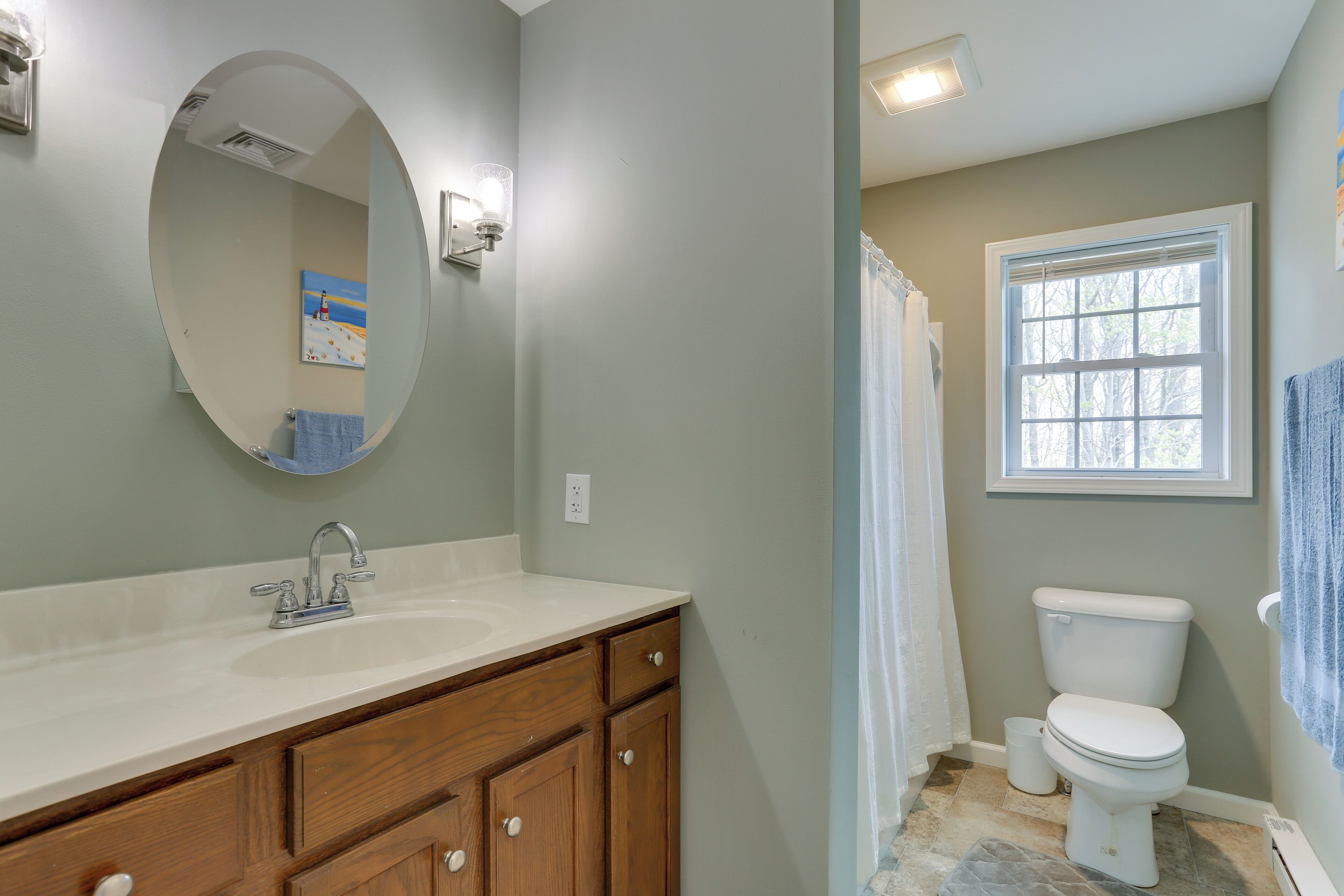 Une salle de bain avec un lavabo blanc, un meuble-lavabo en bois, un miroir rond, des toilettes blanches et une fenêtre donnant sur des arbres.
