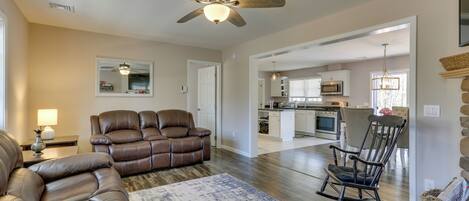 A living room with a brown leather sofa, a rocking chair, a ceiling fan, and a kitchen in the background.