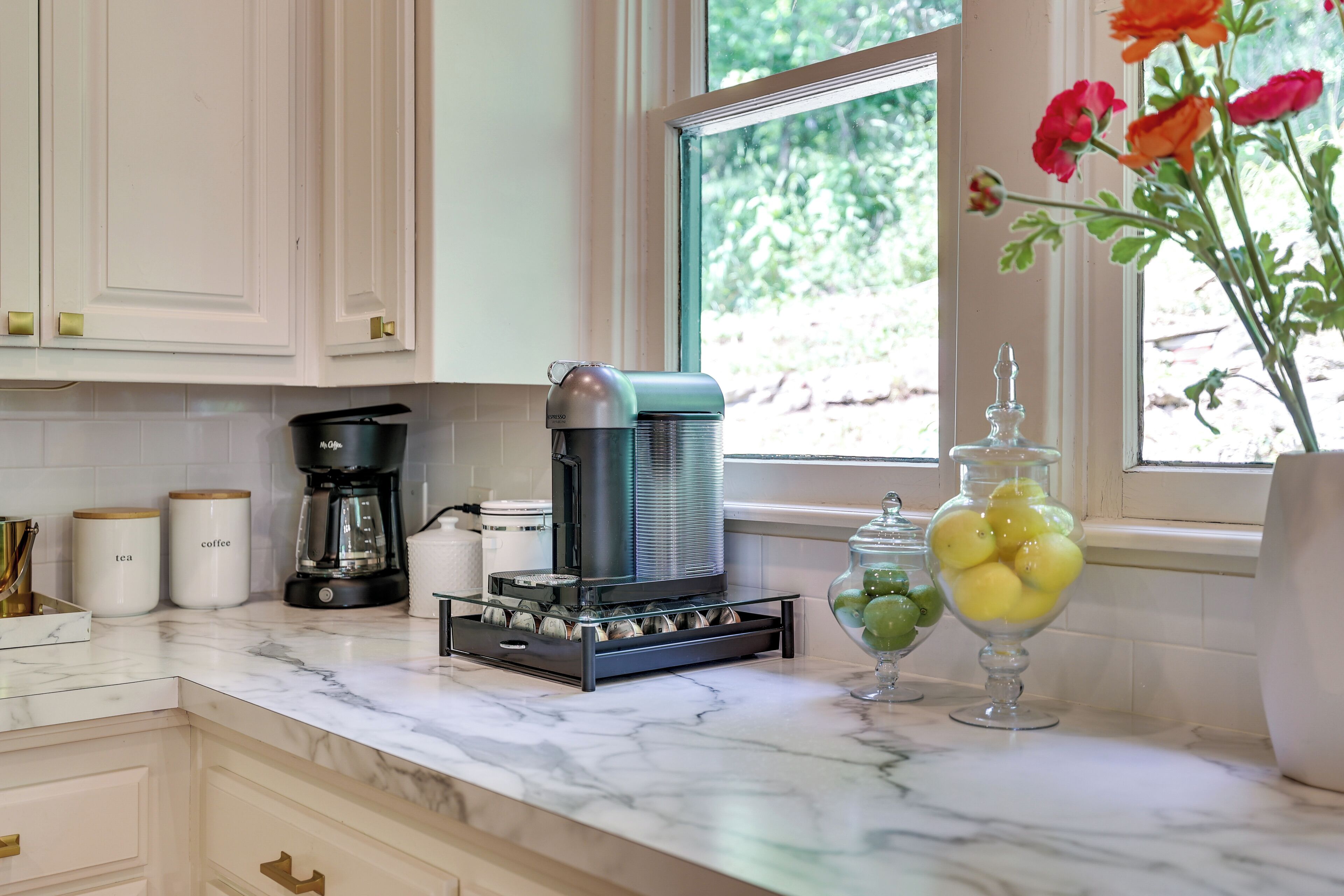 A kitchen with a coffee maker, tea and coffee containers, and a window with flowers.