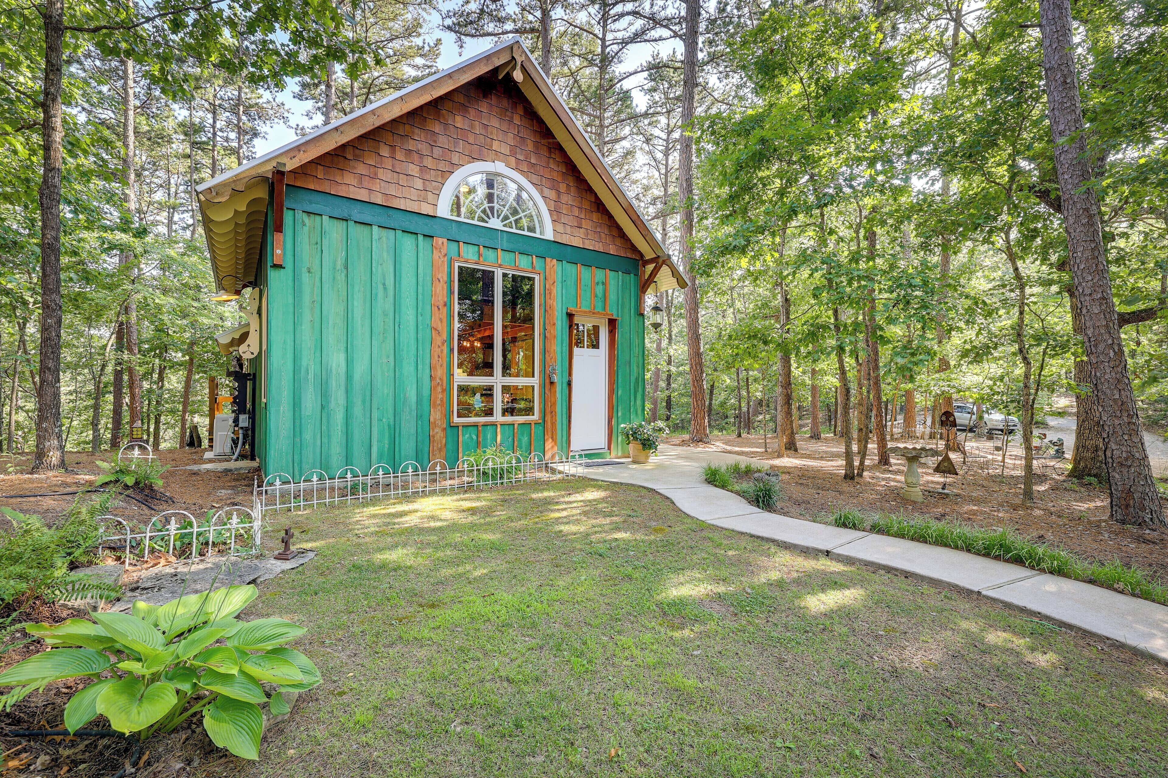 A small, rustic cabin with a green exterior and a large window, surrounded by a forested area.