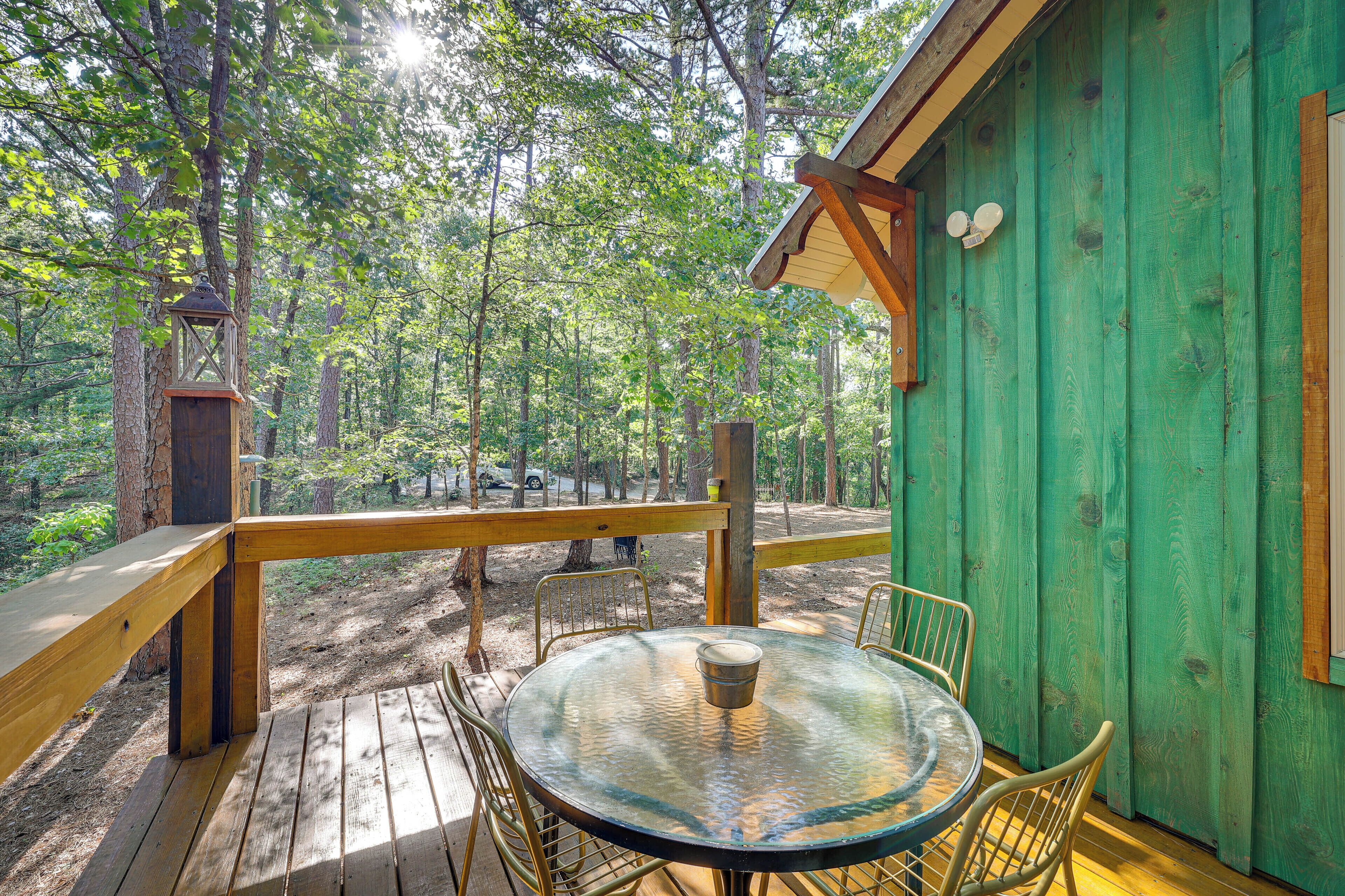 A wooden deck with a glass table and chairs, surrounded by a forested area.