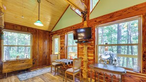 A log cabin interior with a dining area, a television mounted on a wooden pillar, and large windows offering a forest view. - Pet-friendly Eureka Springs Rental w/ Private Deck (Eureka Springs)