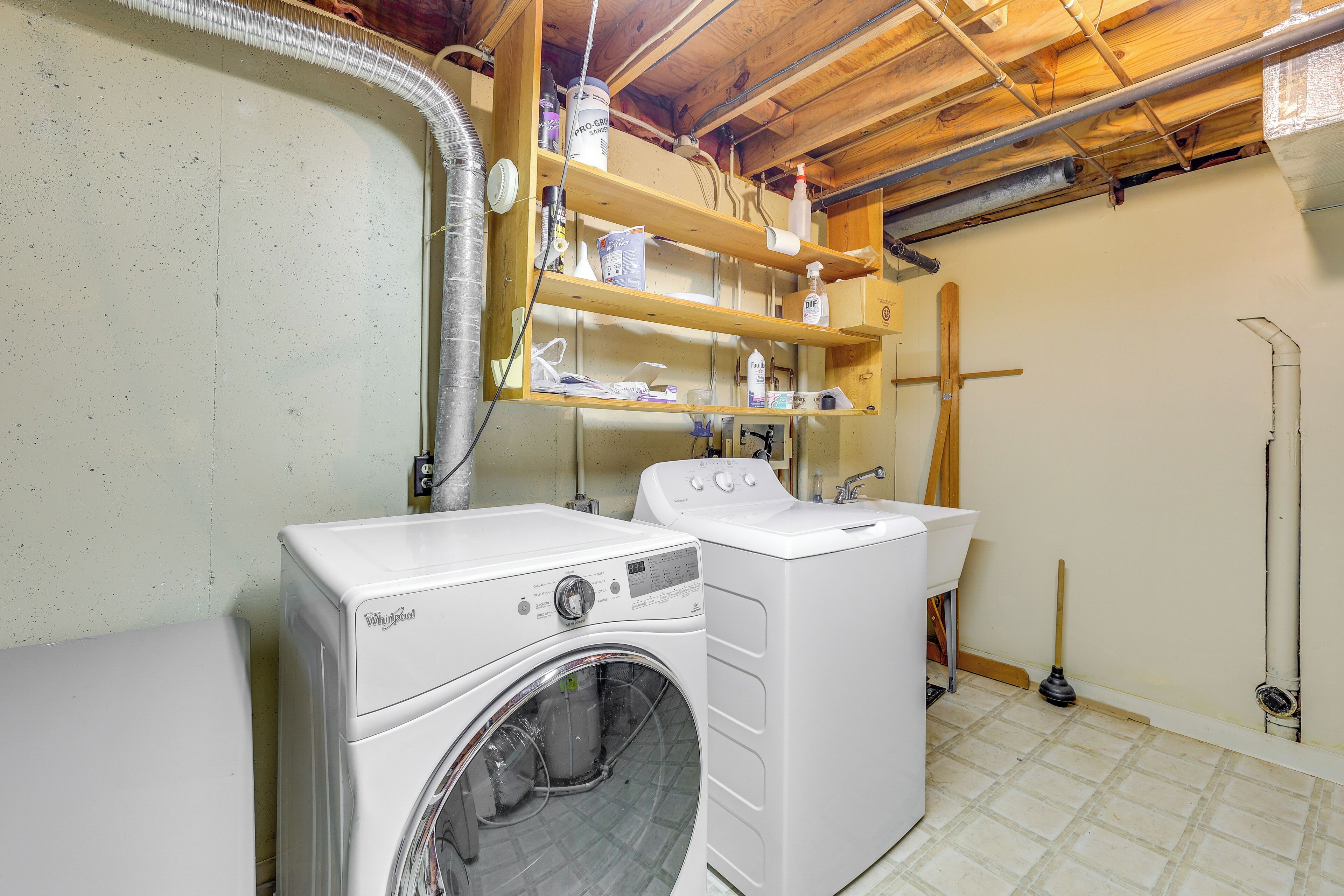 Uma lavanderia com duas máquinas de lavar Whirlpool, prateleiras de madeira acima e um esfregão no chão.